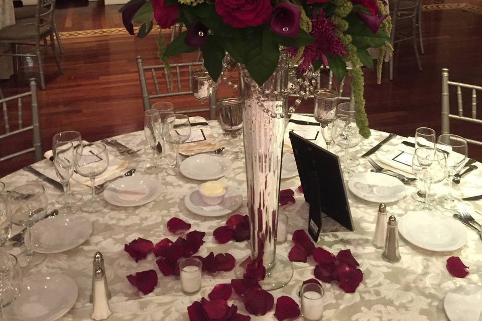 Wedding arrangement designed with black magic roses,red callas , green hanging amaranthus ,and green hydrangeas on top of clear glass pilsner vase .