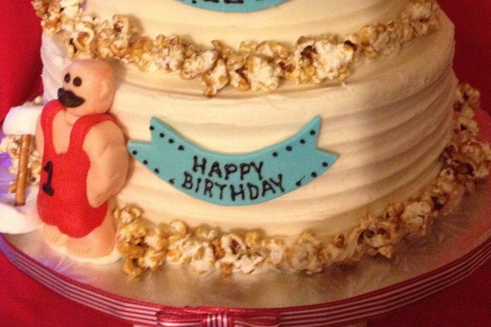 Leo's first birthday cake for his carnival-themed party.  Chocolate cake filled and frosted with sweet American buttercream frosting, decorated with a sugar paste Strong Man and Leo the Lion, garnished with homemade caramel corn.