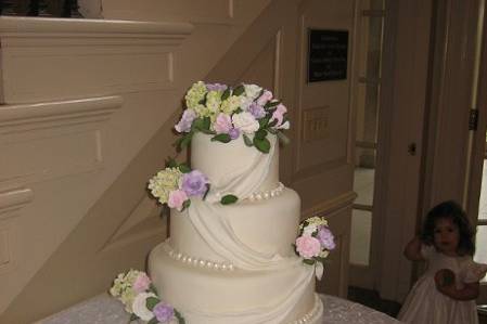 Cake is covered in fondant with fondant draping down each tier. Cake has clusters of sugar roses and hydrangeas