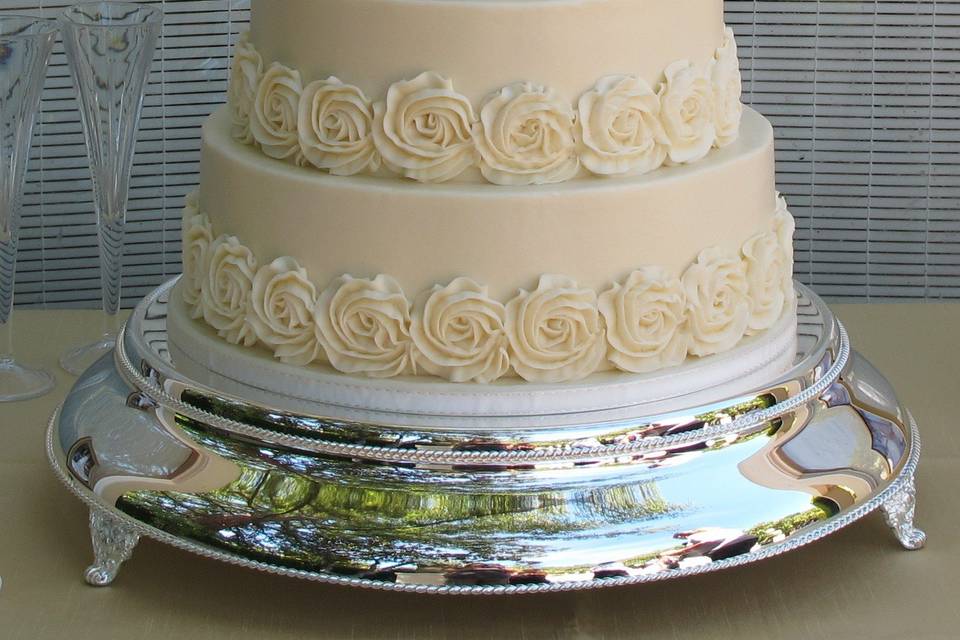 Four tiers covered in smooth American Butter Cream and adorned with butter cream ruffled rosettes to match the bride's gown. Topper provided by the bride and groom.  Displayed on our gorgeous silver cake plateau.