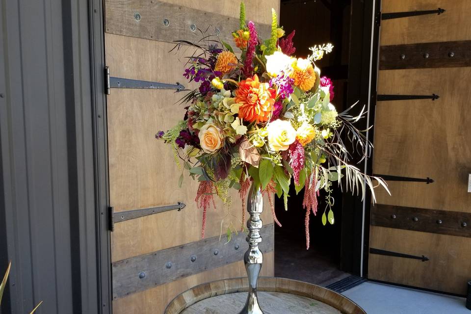 Wine barrel floral, silver riser with a spray of bright vibrant summer flowers including roses, dahlias, stock, ranunculus and greens.