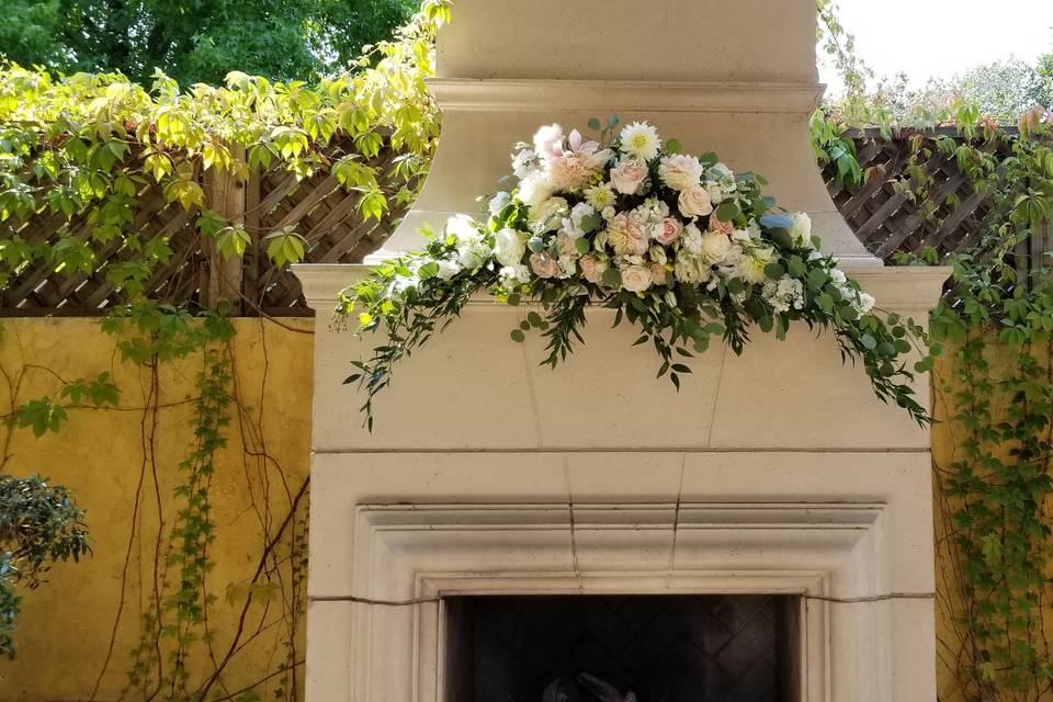 Outdoor fireplace ceremony, mantle flowers include blush and white roses, ranunculus and dahlia with ruscas greens.