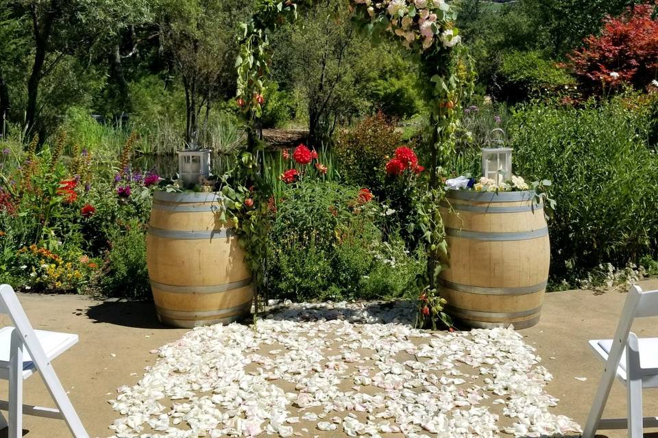 Hans Fahden ceremony by the pond, rustic wedding arch loaded with greenery and fresh flowers. Barrels topped with rustic lantern and fresh flowers.