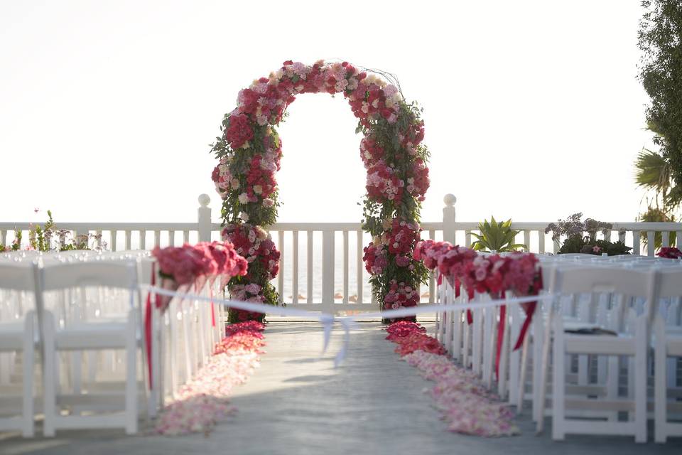 A romantic arch with garden roses, hydrangeas, peonies and many more.  The gradation of colors from hot pink to lighter pinks are reflected by the rose petals down the aisle.  full cones of roses and hydrangeas line the chairs.  Quite and before the wedding.