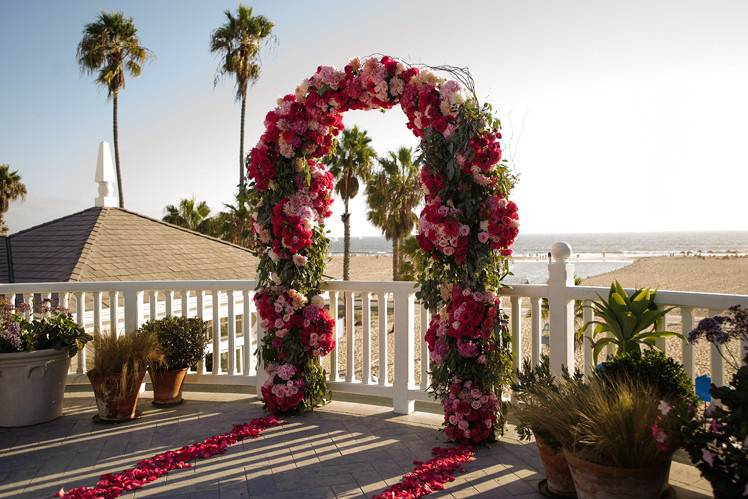 A view of the Santa Monica Beach in the background of the wedding ceremony arch.  This arch is full of garden roses, dahlias, pink hydrangeas and spray roses.