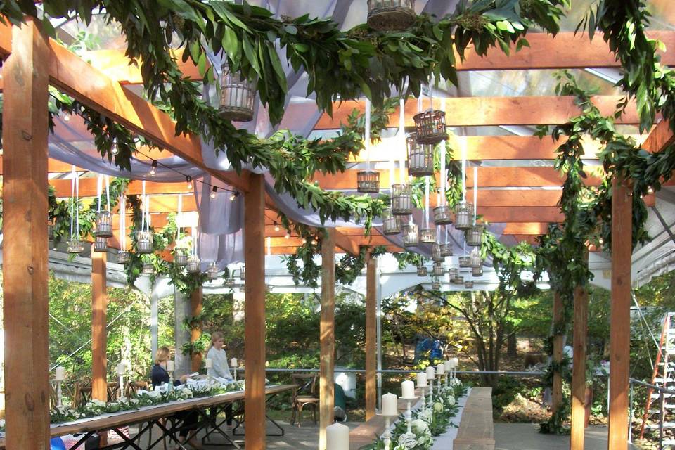 A garden wedding under a custom-built pergola, with
garlands of eucalyptus and ruscus winding through the beams. Long runners of foliages and flowers grace the middle of the table, with candles interspersed. Lanterns also hang from the beams holding the garlands.  Magic!