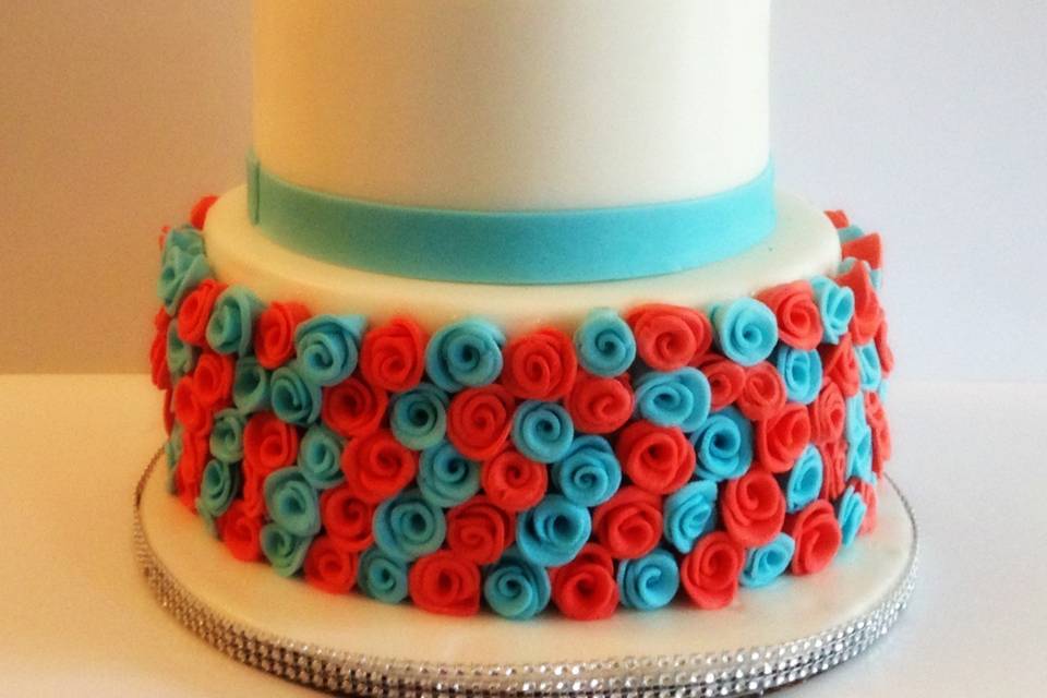 Three tier wedding cake, fondant covered. Bottom tier has red and blue roses going around the tier. Top tier has  a rose bouquet with red and blue borders on the other two tier.