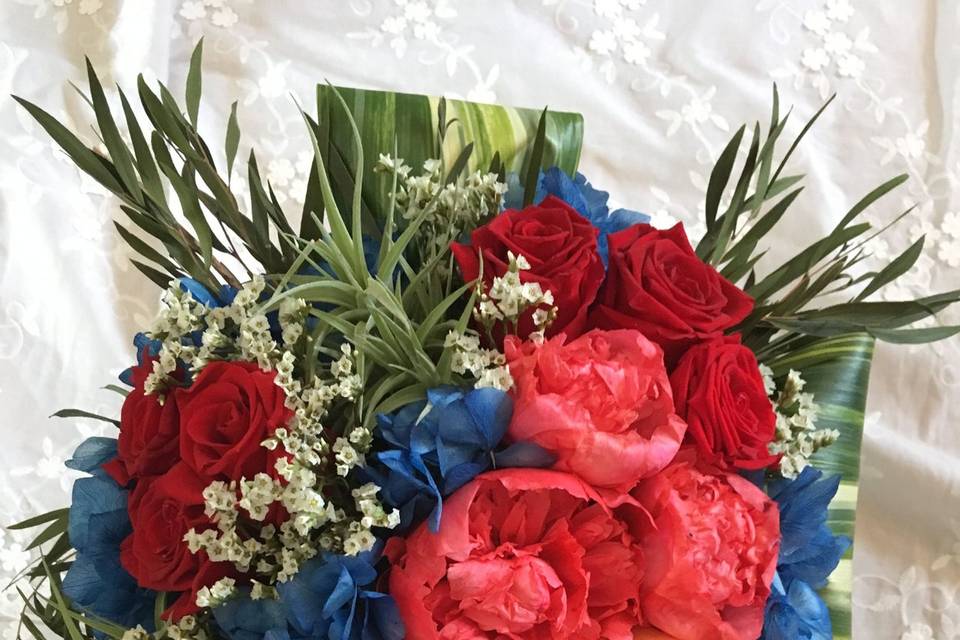 Hand Tied BouquetFeaturing preserved red roses, blue hydrangea, orange callas, and coral peonies. Uniquely featuring air fern plants