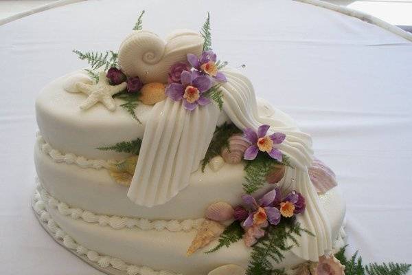 Haley's Orchid Cake features oval tiers that are stacked in a step fashion.  Handmade sugar paste orchids cascade down the 3 tiers, over fondant drapes. Pearlized, edible white chocolate seashells are tucked in the orchids and a large Nautilus shell serves as the cake topper.