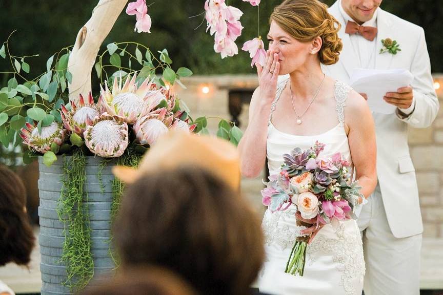 Bride blowing kiss during ceremony, holding succulent bouquet. Hanging orchid altar piece and succulent boutonniere in background.Photo by Jake Holt Photography.