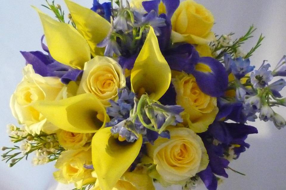 Yellow mini callas, purple iris, light blue delphinium, white waxflower and yellow limoncella spray roses are featured here.