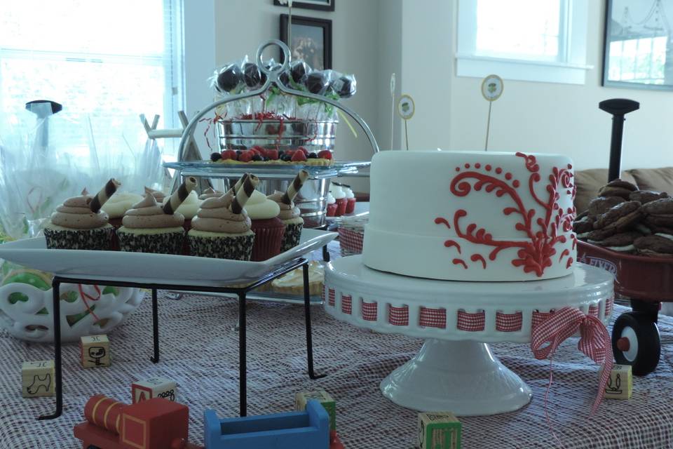 Assorted cupcakes,fondant covered cake with red piped scroll work and cake pops in the background