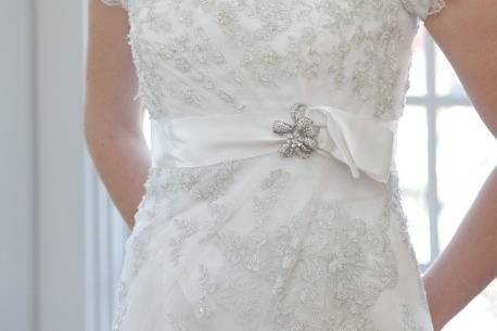 This slightly fitted sheath style gown is created in lace applique. A unique satin band at the empire waist features a lovely silver broach.
Gown features a split on the side as lace drapes down the hem. Lace on netting over organza skirt.