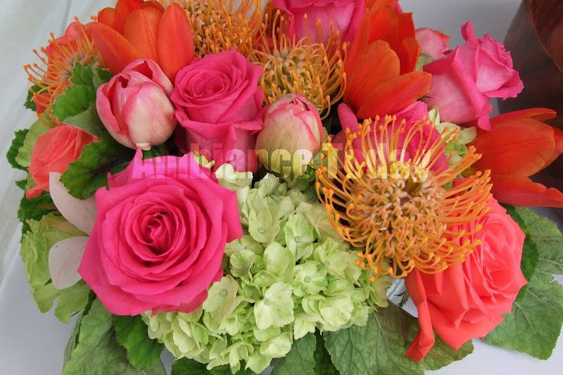 Low, round, bright centerpiece with tulips, pincushion protea, roses, hydrangea and scented geraniums.