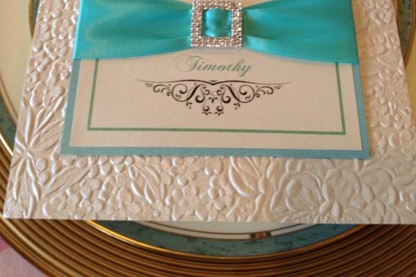 Square embossed floral bi-fold with inset on 3 layers centered on the cover, tiffany blue ribbon with a double square rhinestone buckle.