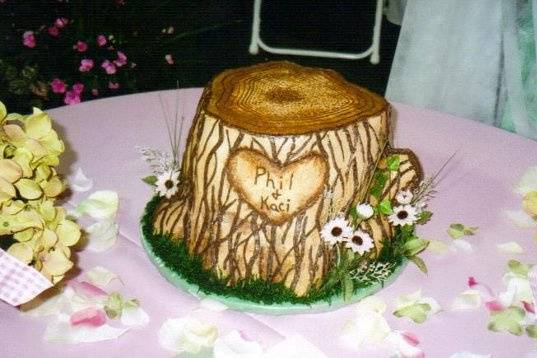 ETCHED IN LOVE:  This cake captures the essence of true love with a heart surrounding the lovers names etched into the side of a tree stump.  It is decorated with silk flowers and leaves.  Some decorations may differ according to the availability of the items used.  This cake is actually a 3-tier cake in disguise with an extra small one to the side (12