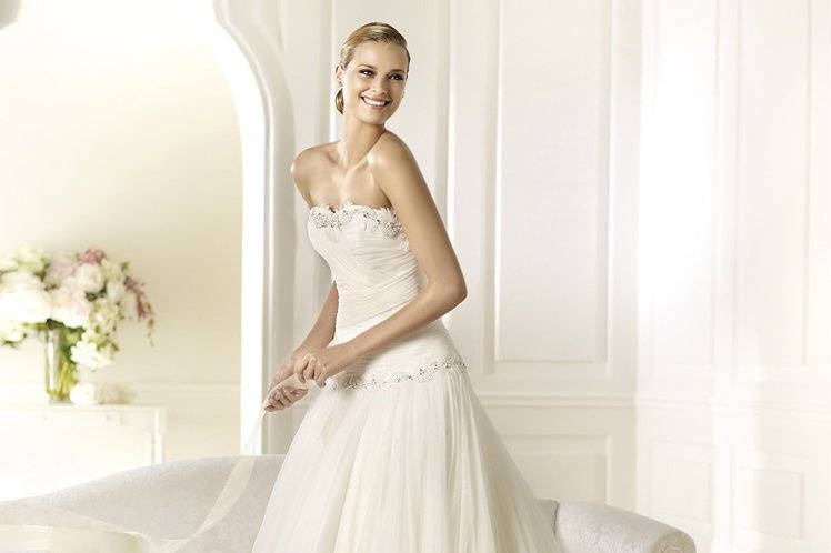 DINAMARCAAll eyes will be on the straight neckline of the Dinamarca wedding dress by Pronovias 2013. Decorated with original gemstone and white feature appliqués, the neckline highlights the beauty of the soft, draped bodice. More of these appliqué details are found around the waistline of this princess wedding dress with a tulle skirt. A softly falling flounce on the skirt gives this wedding dress a marked, modern air.