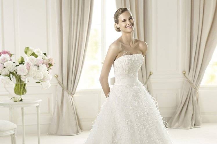 UCIFAUcifa by Pronovias 2013 has a romantic off-the-shoulder neckline. The bodice is made of draped tulle with flower and feather appliqués. This A-line dress has a plain grosgrain belt with a bow at the back, that emphasizes the waist of the flared skirt with its cascading appliqués.