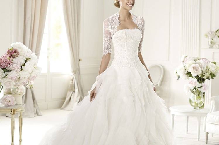 ULUAThere is romance in each and every detail of Ulua wedding dress by Pronovias 2013. This tulle and organza off-the-shoulder design has a Chantilly lace bodice and a jacket with sleeves in the same fabric. This princess cut dress has a skirt with circular frills and is close fitting to the hip.