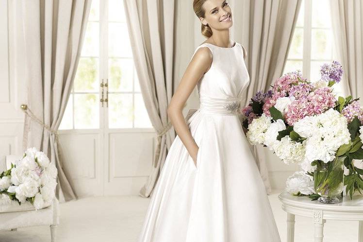 DALILAThe Dalila model by Pronovias 2013 is an unforgettable choice. Mikado, an excellent, soft type of silk, has been used to create this wedding dress with a flattering bateau neckline and a sash that encircles the waist. The delicately pleated sash features a spectacular floral gemstone embroidered brooch. Lateral folds of material flow out from the sides of the dress, hiding some convenient pockets. The final, elegant detail is a long removable train embellished with large bows.