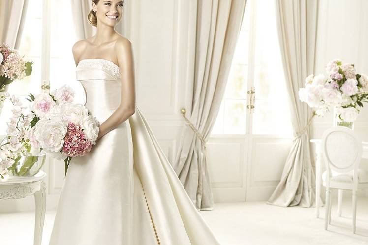 UBINAThe elegant Urbina model from the Costura by Pronovias 2013 collection is made in smooth mikado silk. This A-line wedding dress has a flattering off-the-shoulder neckline with double pleating and soutage embroidery. Beading appliqués surround the bodice and the back has a large bow at the top of a removable train.