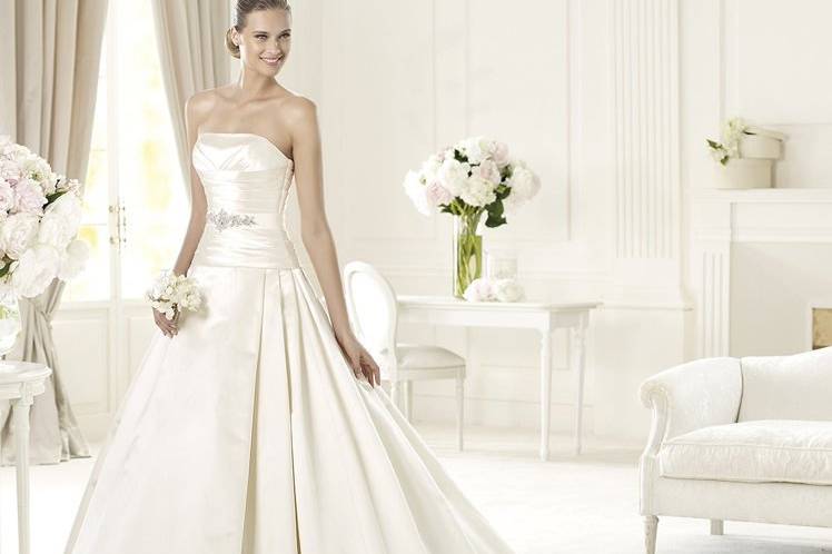 ULLAThe elegant Ulla model from the Costura by Pronovias 2013 collection is made in soft satin. This flattering princess wedding dress has a flattering off-the-shoulder neckline. The waist features an original grosgrain belt decorate with delicate silver beading. The back has a wonderful bow that accentuates the beautiful shape of the dress. The exceedingly elegant skirt has lateral pleats and voluminous train.