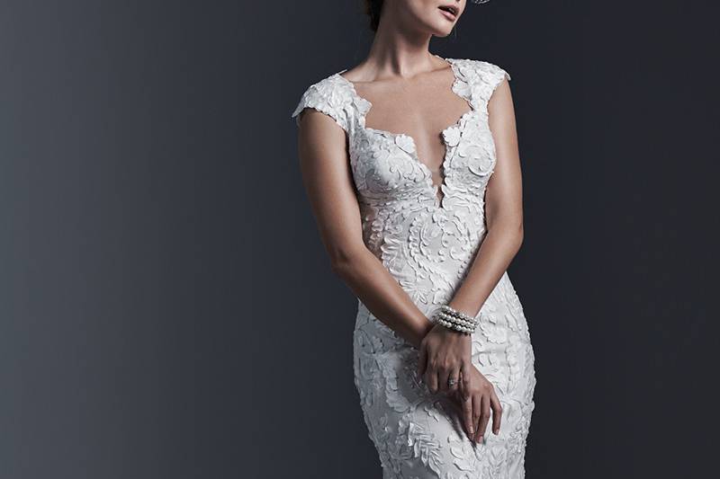 Vidonia <br> Dramatic laser cut lace appliqués lay atop tulle in this form-fitting sheath wedding dress, with demure illusion back. Finished with plunging neckline and covered buttons over zipper closure.