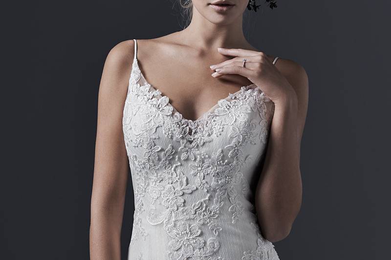 Sandrina <br> Stunning simplicity is found in this embroidered lace and tulle wedding dress with slim A-line skirt. Elegant spaghetti straps give way to a daring, plunging back, edged with lace appliqués. Finished with V-neckline and covered buttons over zipper closure.