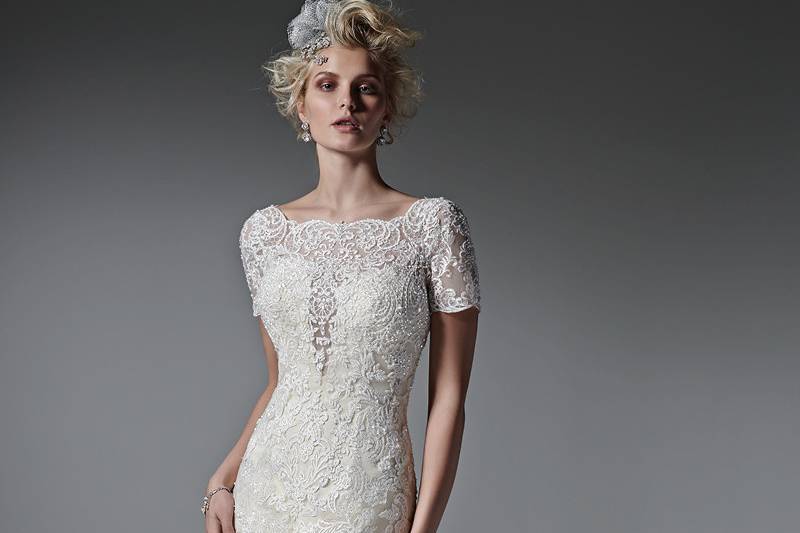 Style Tierney <br> A plunging illusion neckline and delicate, illusion couture length sleeves add a dose of drama to this fit and flare wedding dress, with beaded lace appliqués and a dramatic, open back. Finished with crystal buttons over zipper closure.