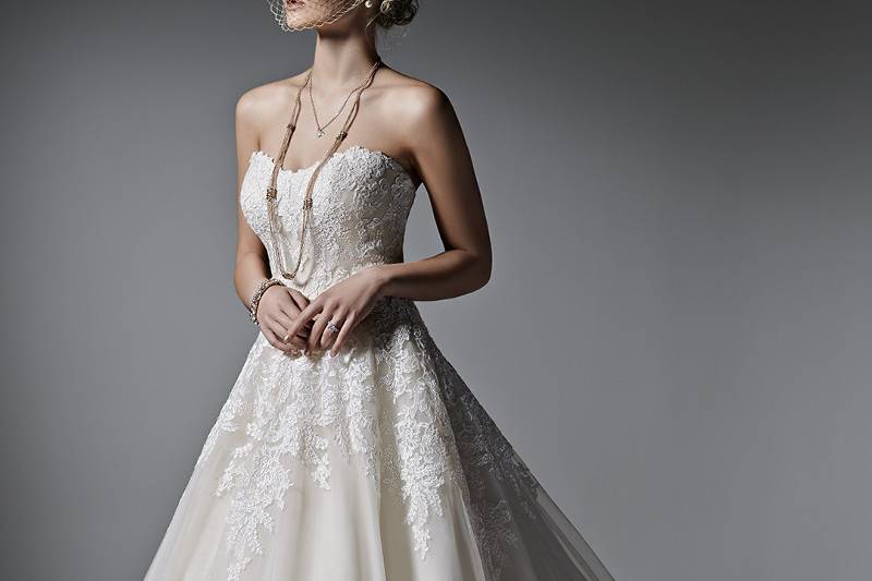 Style Zelinda <br> This feminine ball gown features stunning lace appliqués atop ethereal tulle. A subtle scoop neckline, edged in lace, completes the look. Finished with covered buttons over zipper and inner elastic closure. Lace cap-sleeves sold separately.