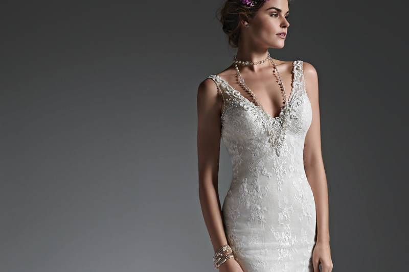 Style Perri <br> Subtle and elegant, this lace sheath wedding dress features a dramatic V-neckline, encrusted with decadent pearls, Swarovski crystals, and glass beads, tracing the tank shoulder straps, and leading down a daring, open back. Finished with covered buttons over zipper closure.
