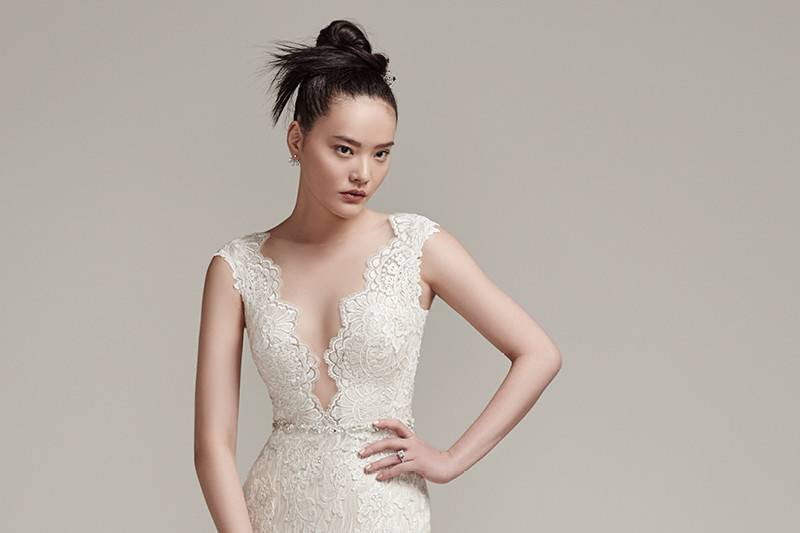 Wyatt <br> Sophisticated and effortlessly glamorous, this modern lace sheath wedding dress flaunts a sexy, plunging illusion neckline, beaded belt, and lace cap-sleeves. Complete with illusion back with ruched details. Finished with pearl buttons over zipper closure.